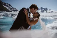 Bride and groom share their first kiss beside brilliant blue glacial water, surrounded by snow-covered Alaska mountains.