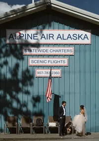 Bride and groom stand outside the Alpine Air Alaska hangar beneath signs advertising statewide charters and scenic flights.