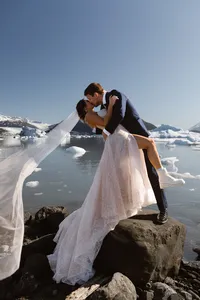 Groom dips and kisses the bride on a rocky shore, her veil flowing beside an Alaska glacial lake filled with icebergs.