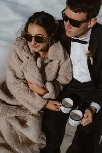 Newlyweds sit close together during their glacier picnic, holding matching mugs while the bride bundles up in a cozy coat.