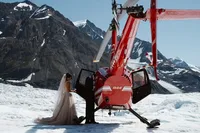 A bride in a flowing white wedding dress and long veil stands beside a red helicopter on an Alaskan glacier as the groom opens the door, surrounded by dramatic snow-covered mountains.