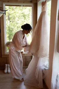Bride in pale pink pajamas reaches for her lace wedding dress hanging beside a sunlit cabin window.