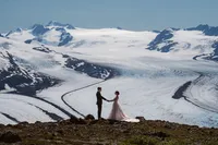 Newlyweds hold hands on a rocky ridge, dwarfed by a vast snow-covered Alaska glacier and surrounding mountain peaks.