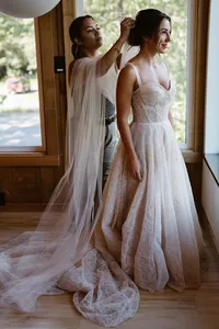 Bride in her lace wedding dress stands by a cabin window as someone arranges her long sheer veil.