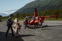 Bride and groom approach a red helicopter on the tarmac, preparing to fly into the Alaska mountains for their elopement.