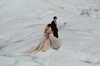 Bride carrying her bouquet walks beside the groom across the vast white surface of an Alaska glacier.