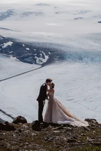 Newlyweds kiss on a rocky ridge with the vast blue-white expanse of an Alaska glacier behind them.