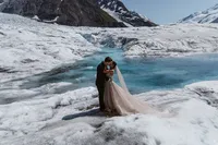 Bride and groom embrace on the glacier beside vivid turquoise meltwater, her long veil trailing across the ice.