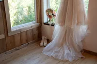 Bride’s lace wedding dress hangs beside a window, with her bouquet and white lace-up boots arranged nearby.