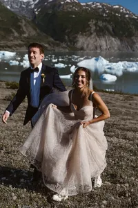 Smiling newlyweds walk along the shore of an Alaska glacial lake, with blue-white icebergs floating behind them.