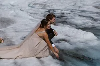 Bride and groom crouch together beside a deep blue crevasse on an Alaska glacier, smiling as they explore the ice.
