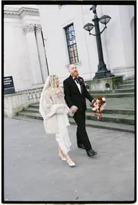 bride with a fur on and groom in black texan suit walking outside marylebone town hall winter wedding photo