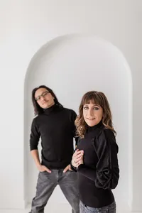 A woman and man stand before a white arched wall, both in black sweaters and jeans. Captured by professional portrait photographers in Washington DC, the woman faces the camera while the man stands slightly behind with hands on his hips.