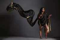 A woman stands confidently in a black and gold saree against a dark background, the fabric billowing dramatically. Barefoot and adorned with jewelry, she smiles slightly—an elegant example of personal branding portraits Washington DC.