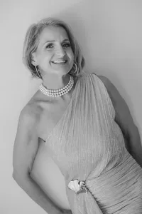 A smiling woman with short hair wears a one-shoulder textured gown, multi-strand pearl necklace, and large hoop earrings. Posing against a plain background, she embodies the elegance of editorial portrait photography in the Washington DC metro area.
