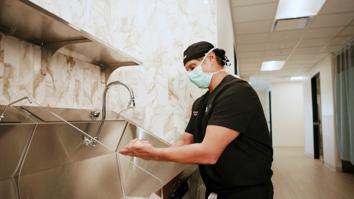 Surgeon in black scrubs and a mask washing his hands at the scrub sink