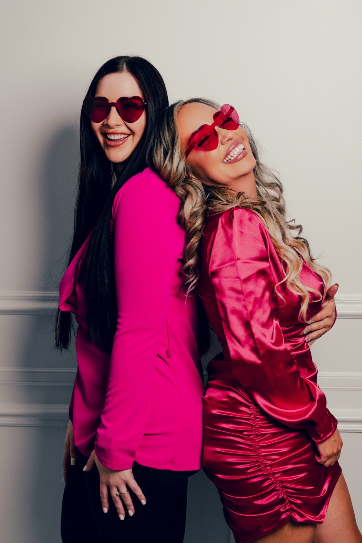 Two women in pink outfits and heart sunglasses posing back to back