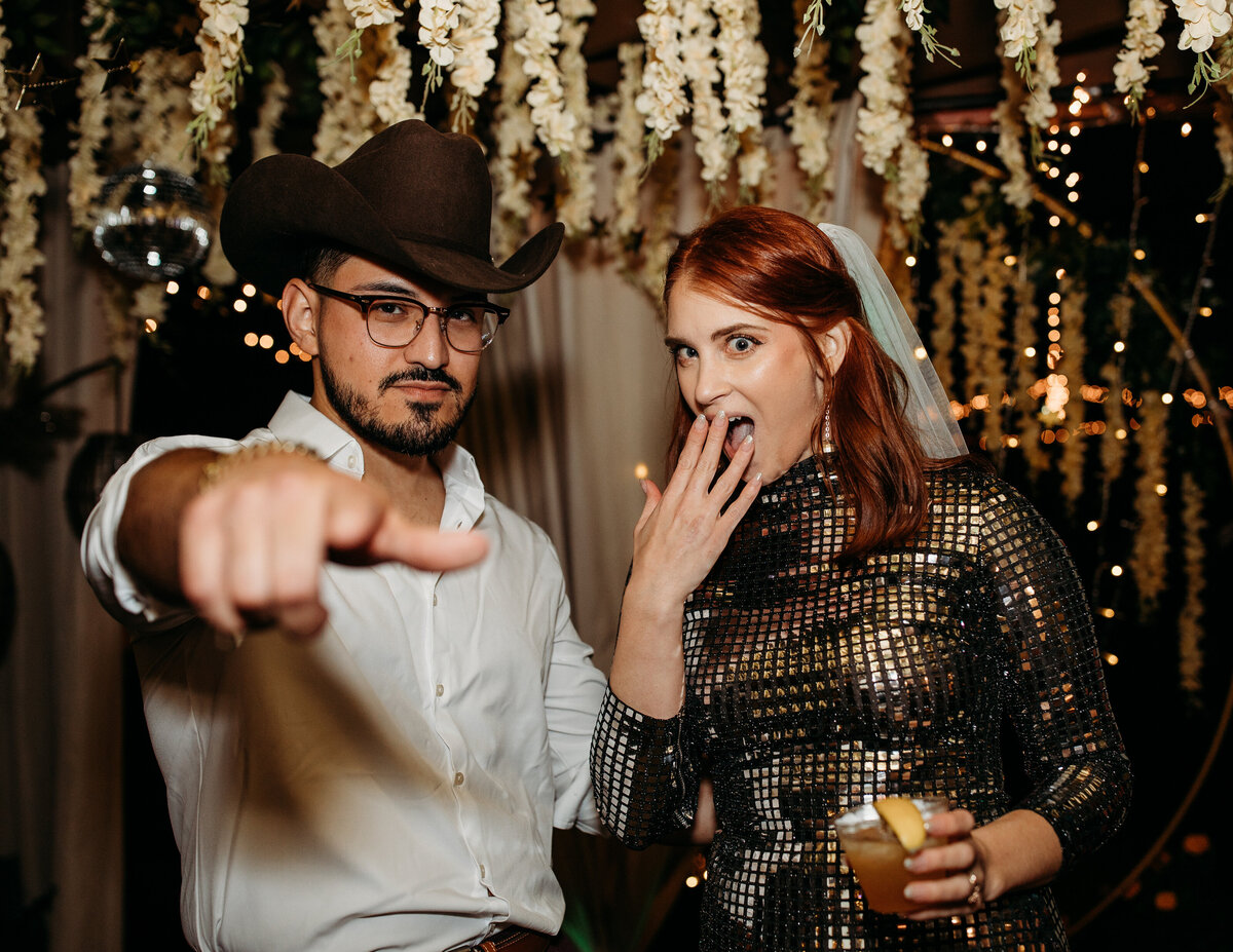 Couple at wedding reception playfully pointing at the camera; man in a cowboy hat and woman with red hair and sparkling dress