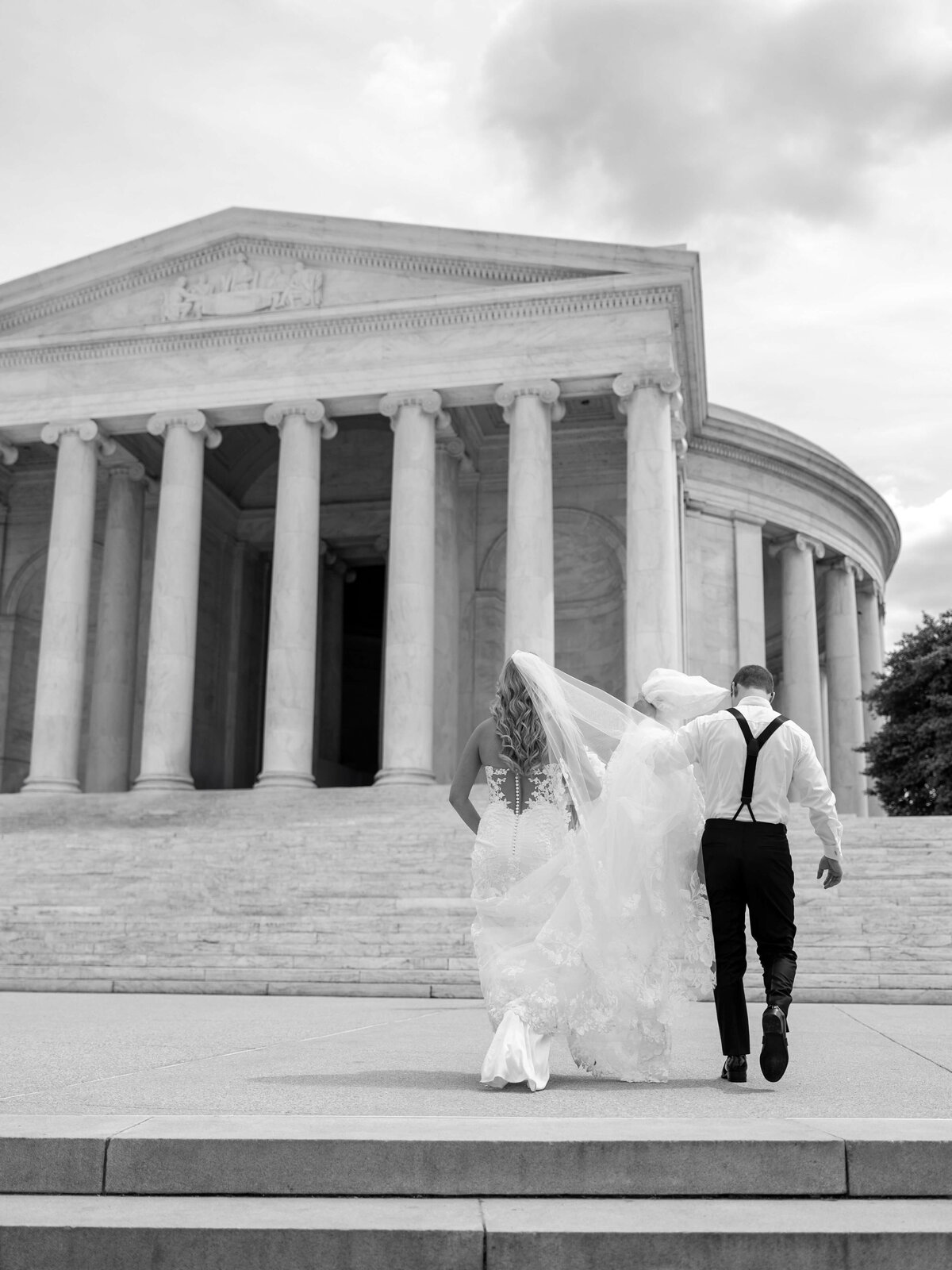 A black and white image of a bride and groom walking hand in hand towards a neoclassical building with large columns. The brides flowing gown and veil are prominent as they ascend the steps together.