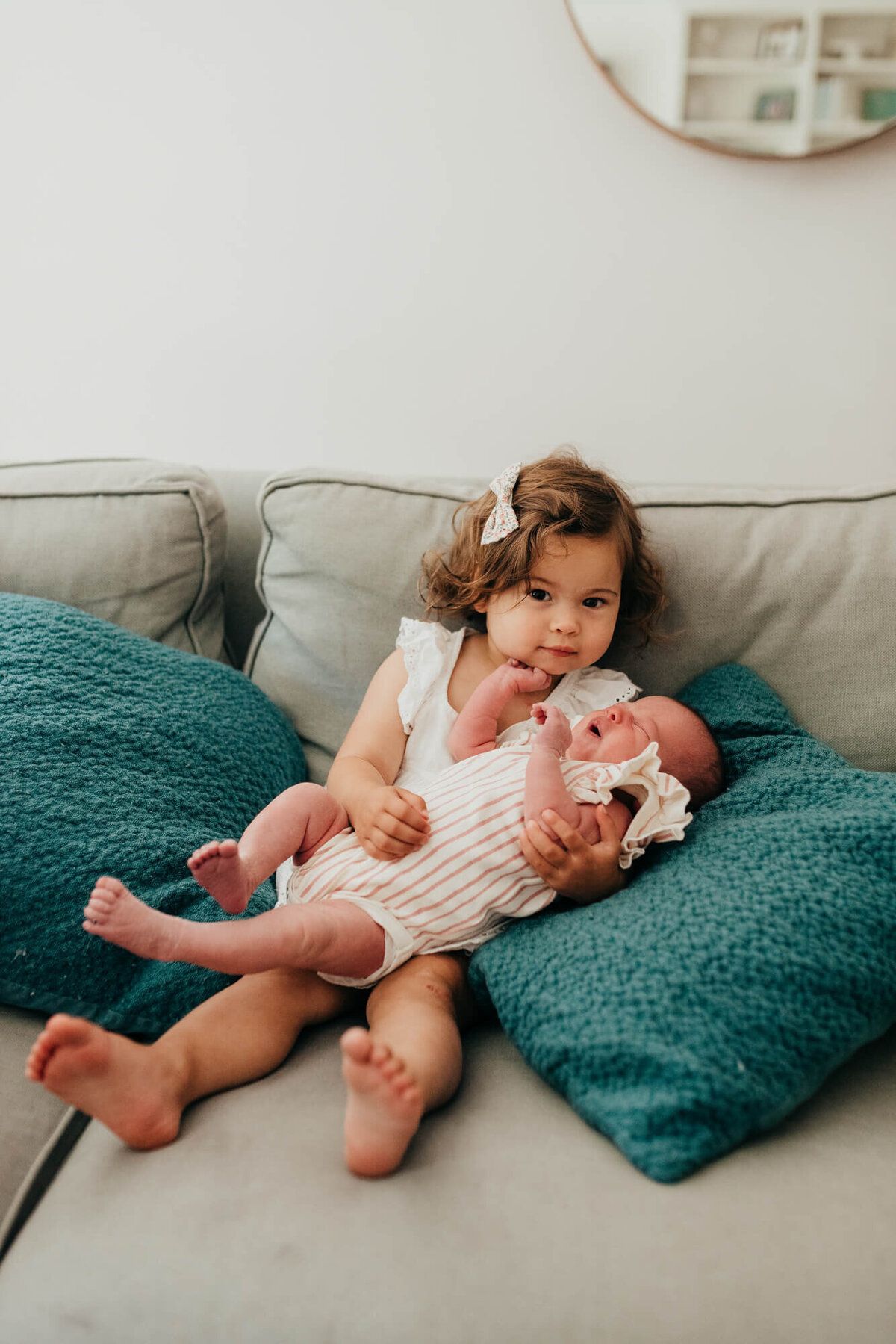 A little girl wearing a white shirt holds her newborn baby sister on the couch