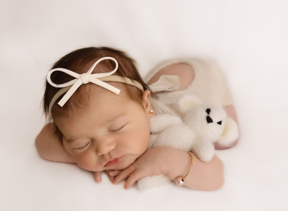 Baby girl is sleeping for her newborn photoshoot. Baby is laying on her belly with her hands folded under her chin. She has a thin bow headband and pearl bracelet and has a felt teddy draped under one arm. Captured by top Brooklyn newborn photographer Chaya Bornstein