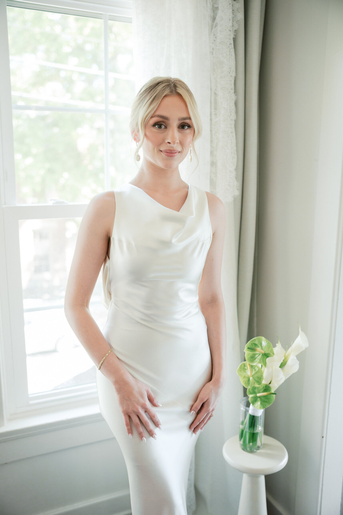 Bride in sleek satin gown poses near window with green floral arrangement, photographed in Dayton by a Philadelphia photographer.