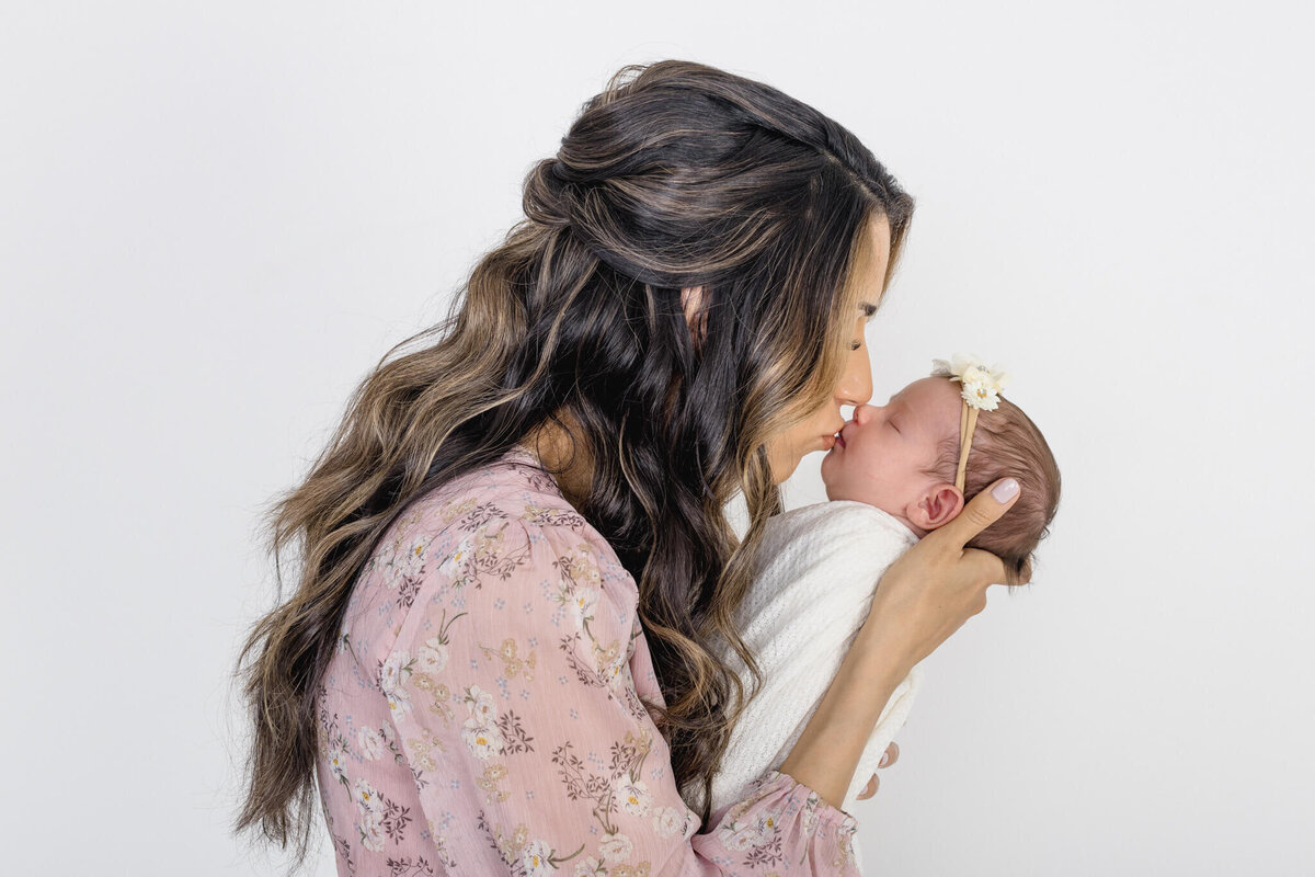 A woman with long, wavy hair gently holds a newborn wrapped in a white blanket, touching noses in a tender moment. Shes wearing a floral-patterned pink blouse, and the baby has a small headband with a flower. The background is plain white.