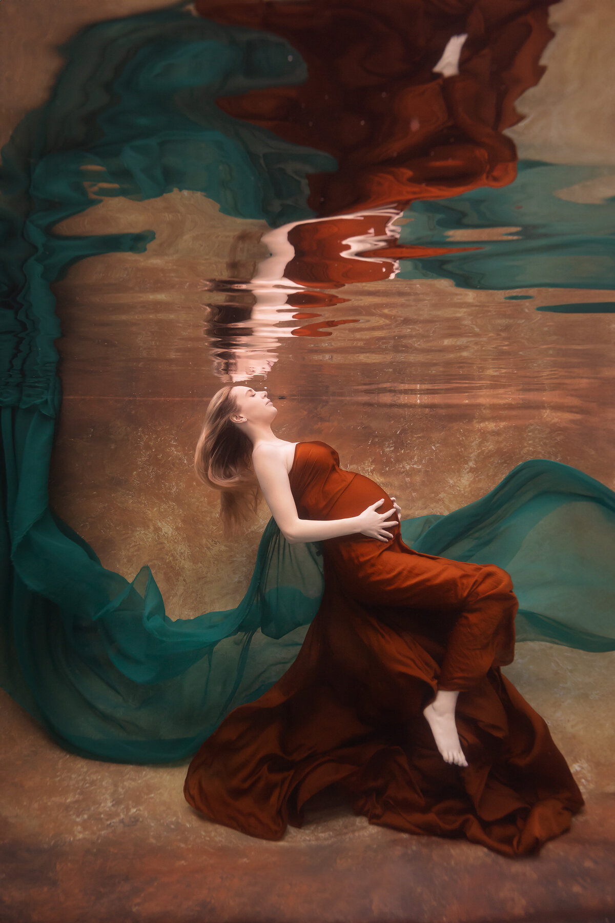 A luxurious underwater portrait by Lindsay Walden Photography, featuring a woman draped in a rust-colored gown with flowing emerald fabric around her. The golden tones of the backdrop and water enhance the ethereal, dreamlike feel of this fine art underwater maternity photoshoot.