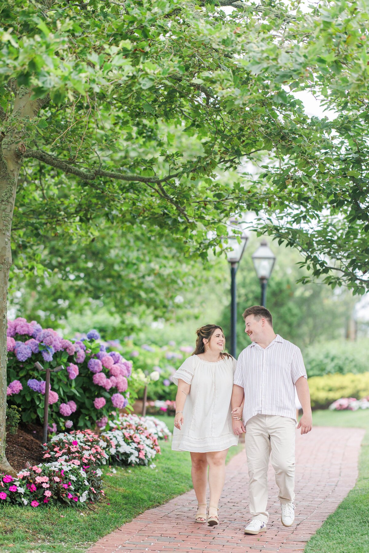 Couple walking hand in hand down a brick path lined with colorful hydrangeas and flower beds, smiling at each other under the shade of leafy trees at Chatham Bars Inn in Chatham, Massachusetts — representing the portfolio of a Cape Cod engagement photographer.
