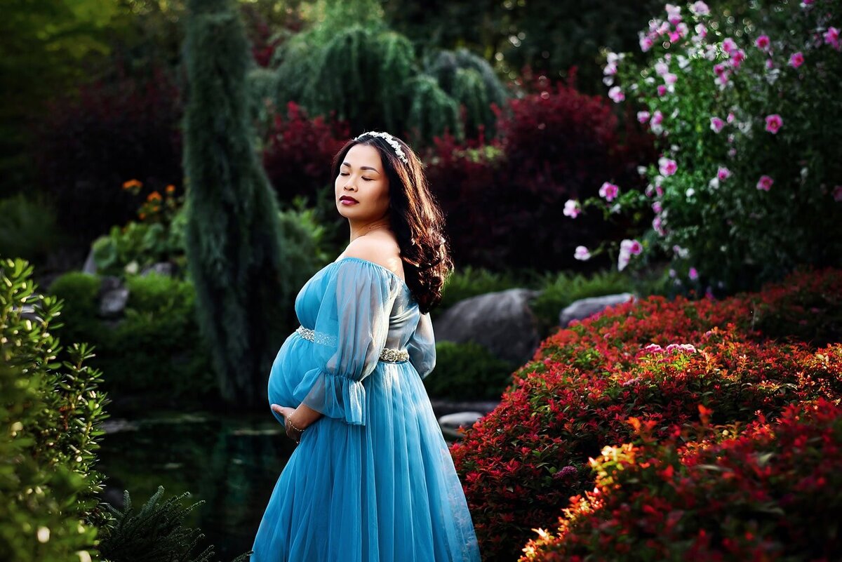 Expecting mother in a blue chiffon dress at Deer Lake Park in Burnaby surrounded by rich, lush foliage for a maternity photo shoot.