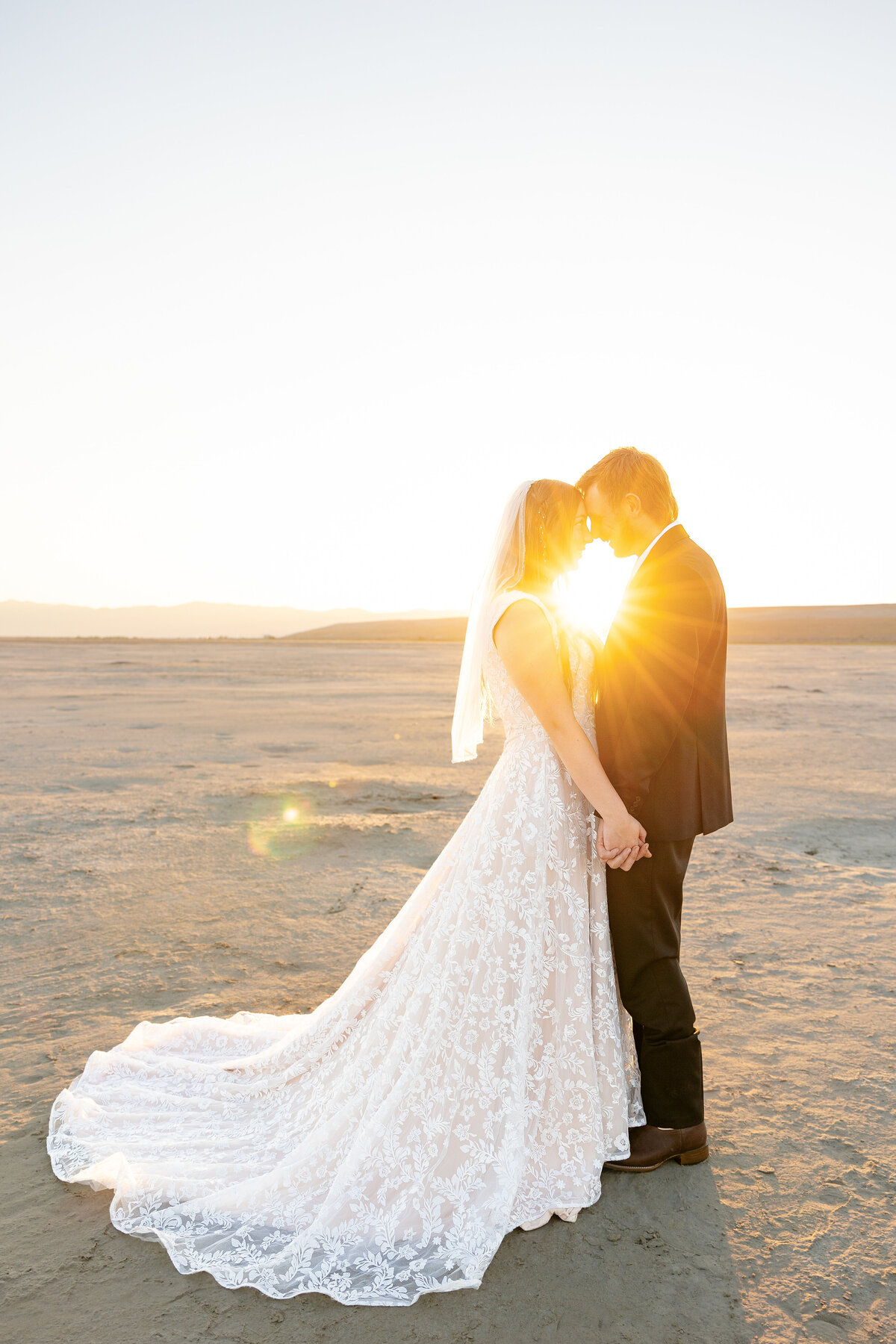 Bride and groom hold hands, foreheads touching, as the golden sunrise glows behind them on the Salt Flats.