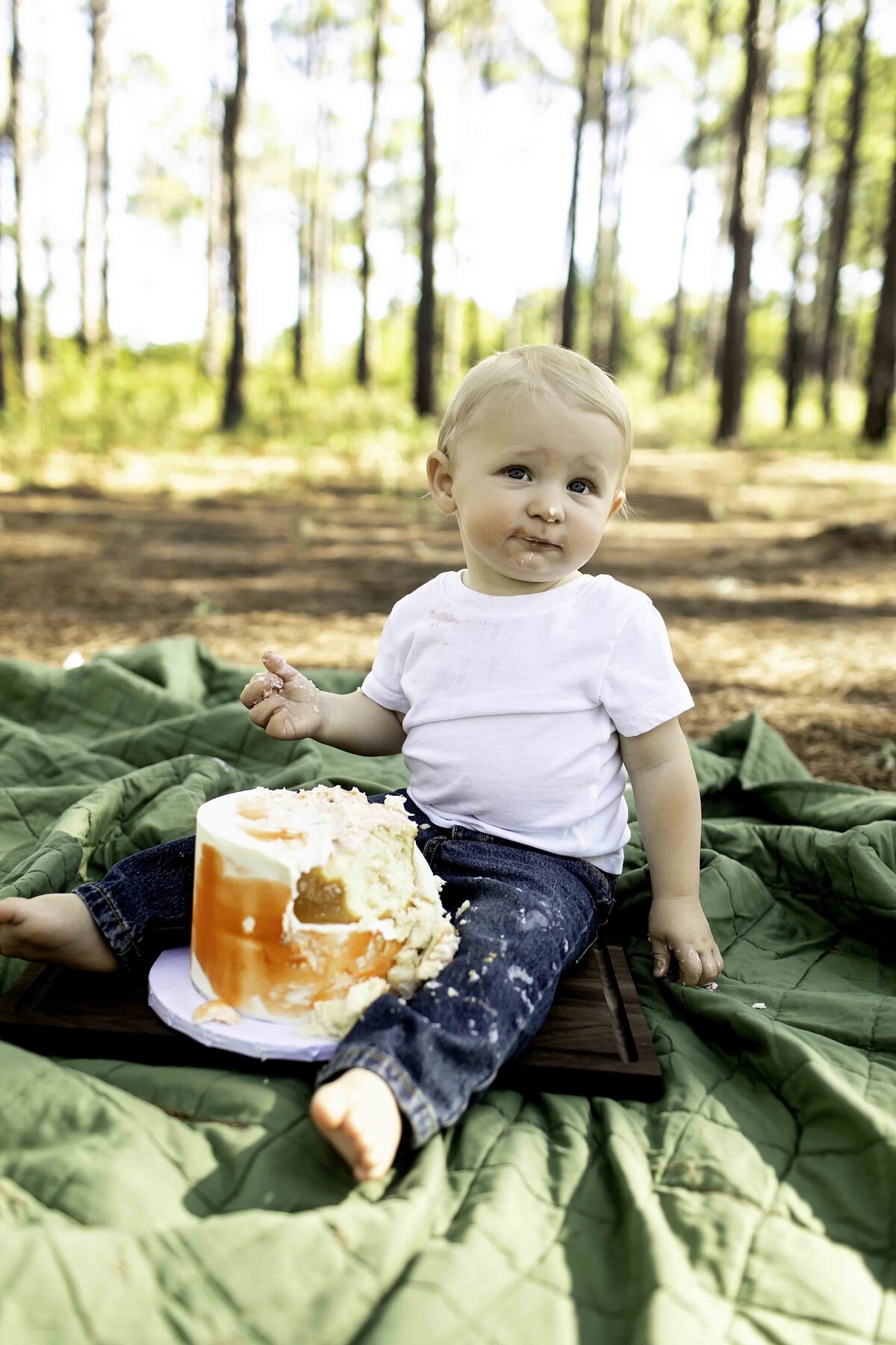 Milestone birthday boy in suspenders and overalls, eagerly smashing his birthday cake while sitting on the grass, with pine trees in the background
