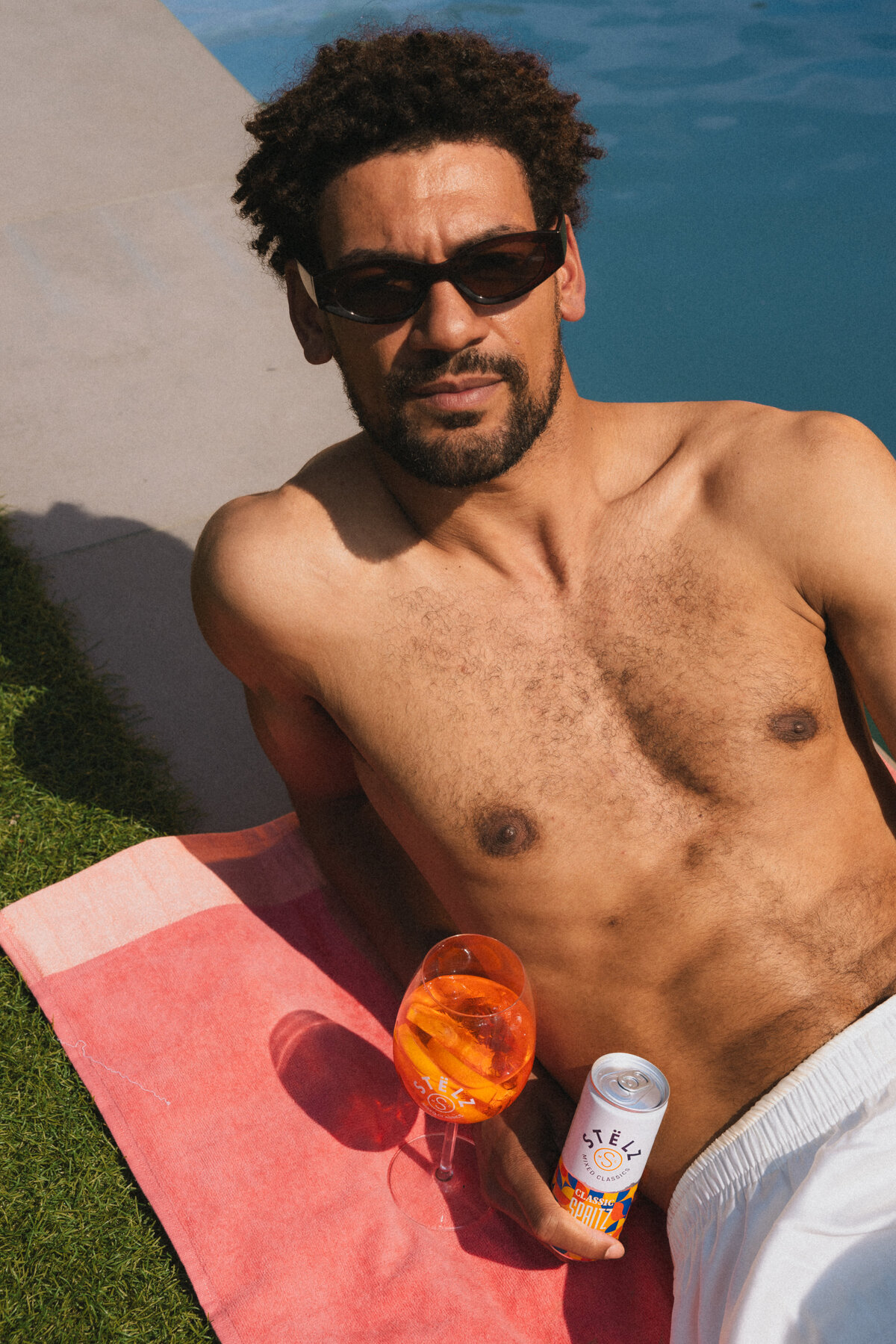 A man casually lies on a towel by the poolside, wearing sunglasses and holding a glass of Aperol Spritz, with a STËLZ can placed next to him on the ground. The sun casts a warm glow on his skin, evoking a strong sense of summer. Photo taken by Bo Eppink.