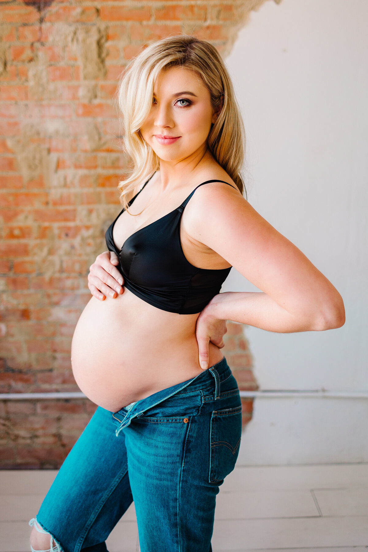 A stunning maternity photo of a pregnant woman with blonde hair, dressed in a chic black bralette and blue jeans. Captured in an Albuquerque photography studio, the image highlights her glowing pregnancy and stylish outfit against a clean and professional studio backdrop.