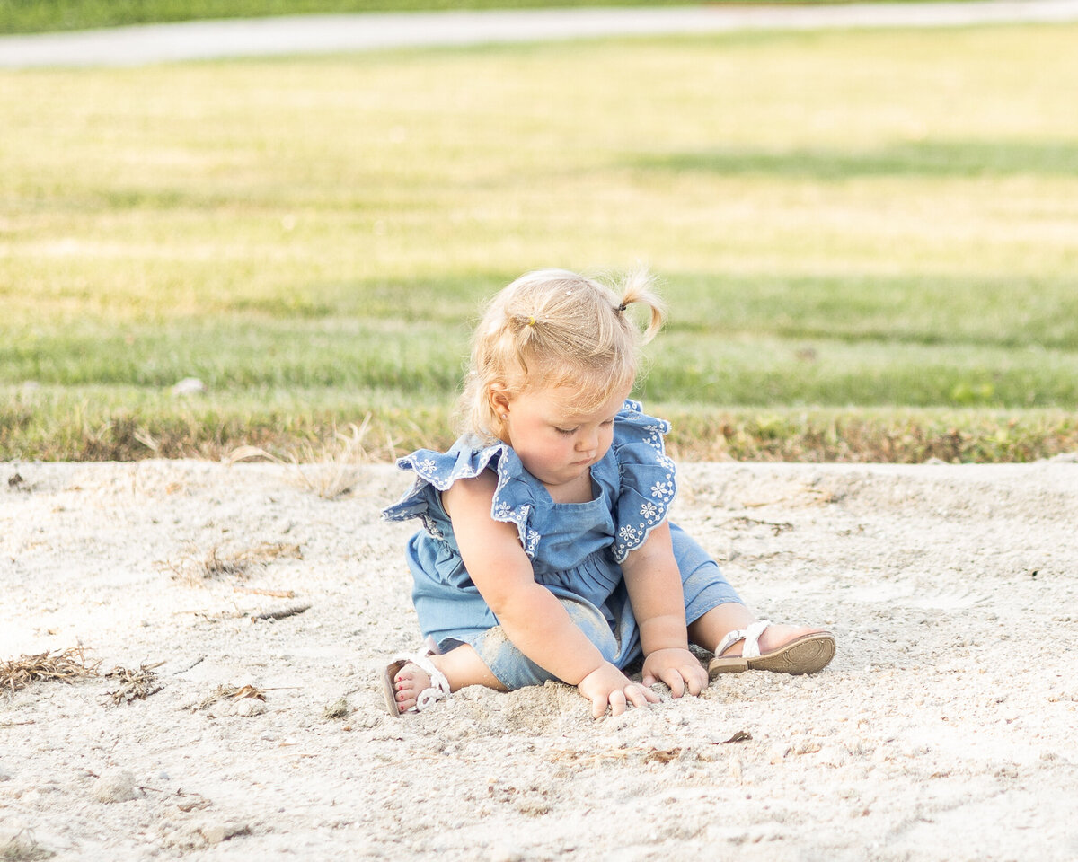 Toddler girl sitting on the ground playing, with blonde pigtails, wearing a light blue dress and white sandals