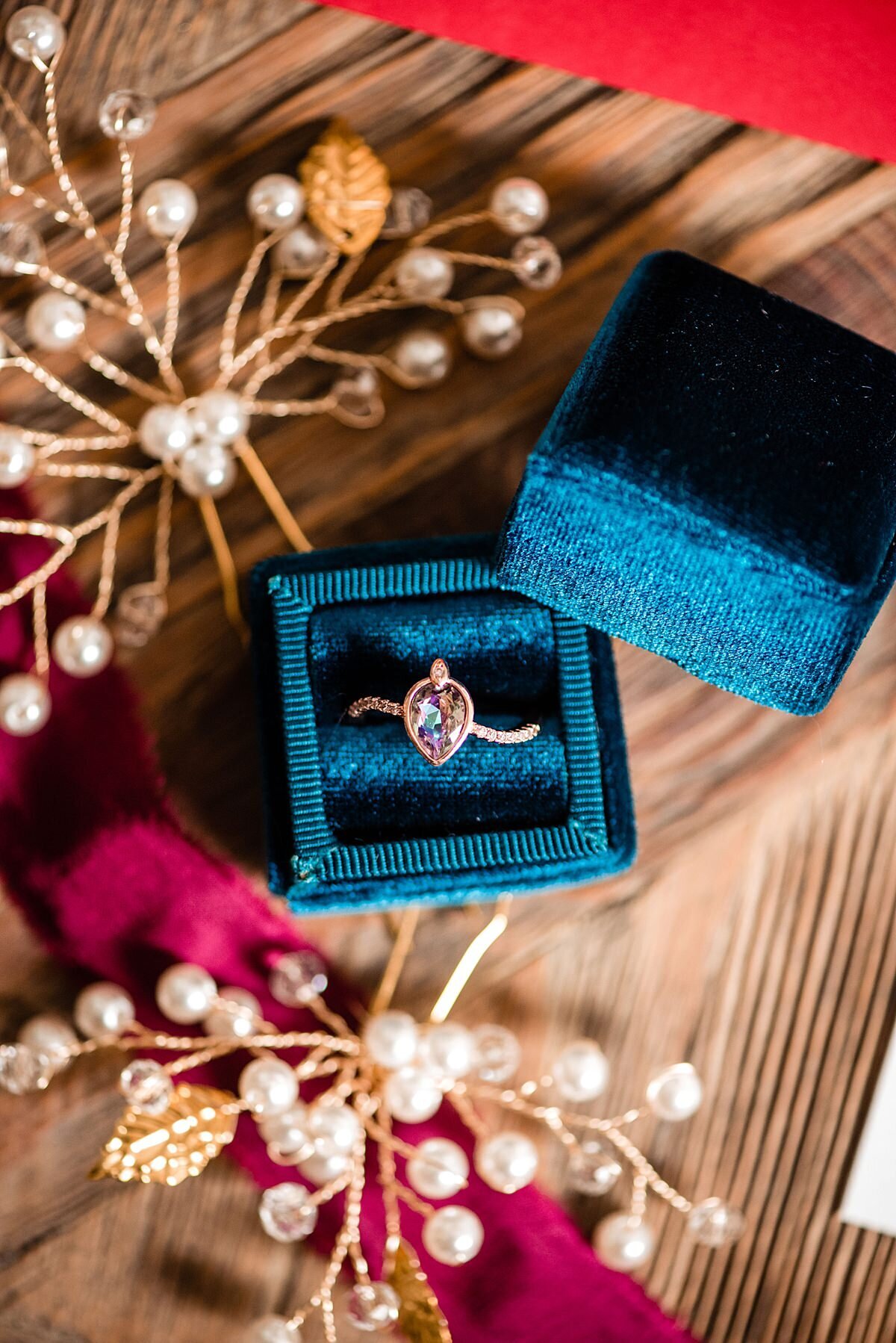 Antique rose gold sapphire engagement ring in a dark teal velvet ring box accented by gold and pearl hair combs with a raw silk burgundy ribbon on a farm table at Arrington Vineyards.