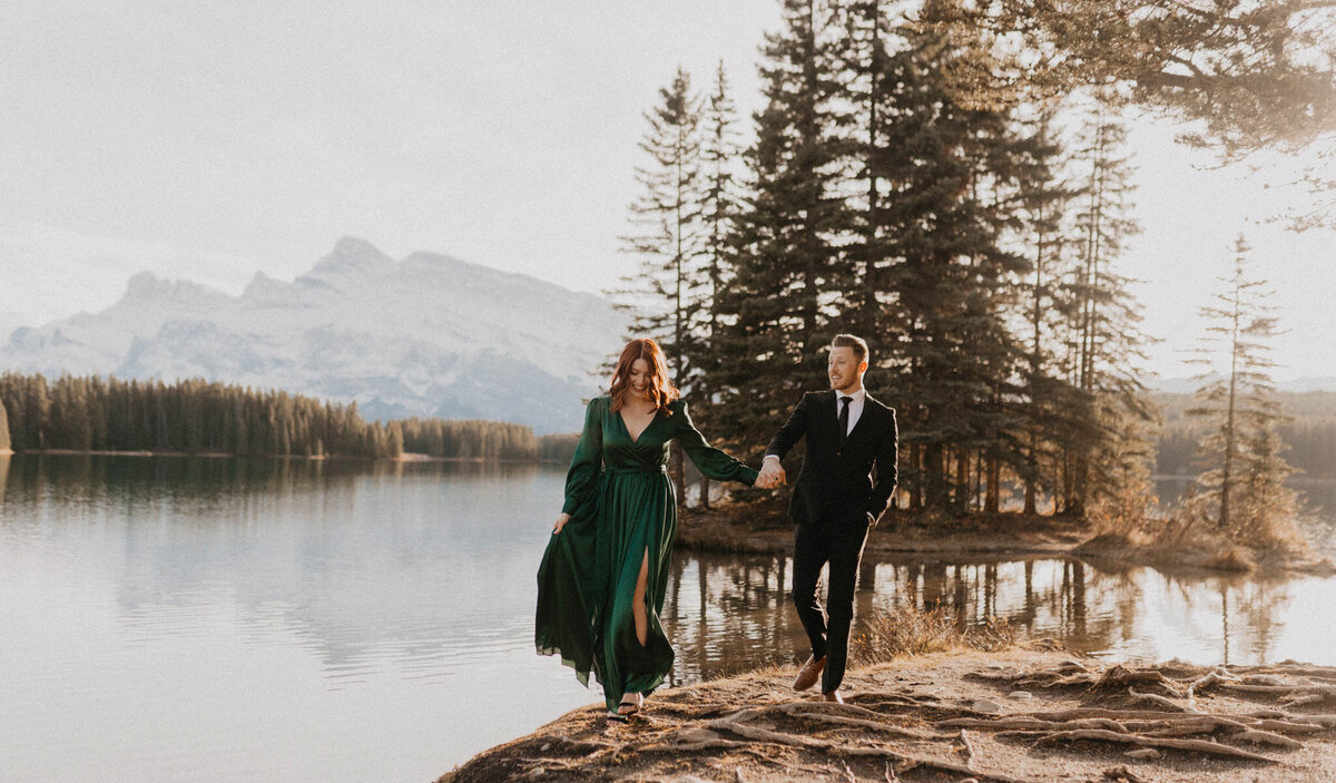 Couple holding hands, walking along the shoreline near water in the mountains, captured by Ash Maclean Photography, romantic elopement and wedding photographer in Red Deer, Alberta. Featured on the Bronte Bride Vendor Guide.