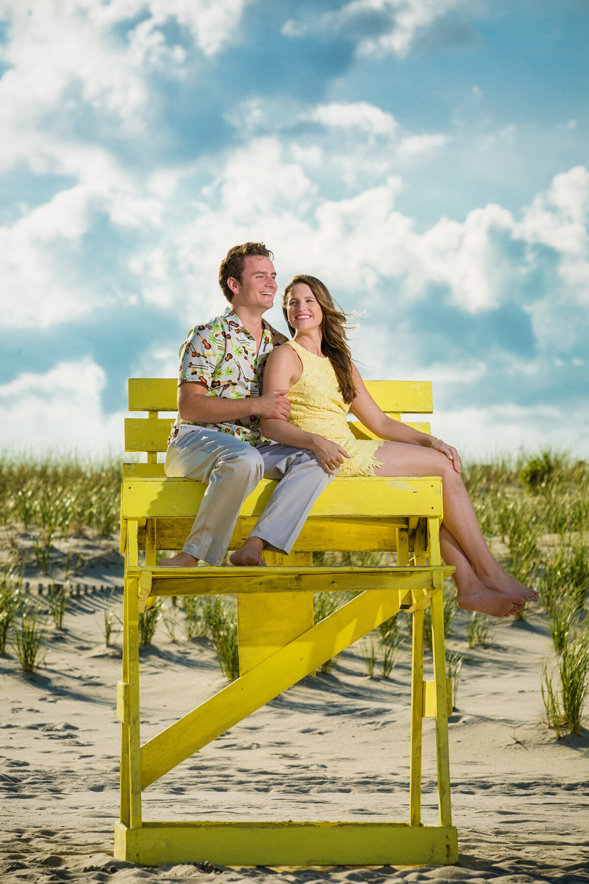 Playful engagement photo of a couple sitting in a lifeguard chair at the New Jersey Shore, with sandy beaches and the sparkling ocean as the backdrop under a sunny sky.
