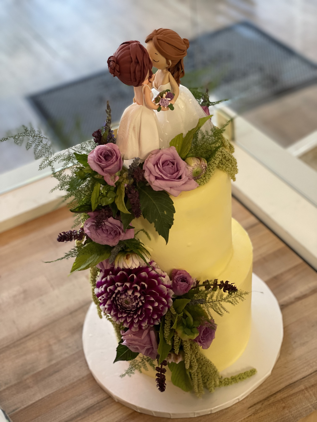 Two-tier gluten-free wedding cake decorated with purple and mauve flowers and a wooden couple topper, handcrafted by Grain Artisan Bakery in Snohomish, WA.
