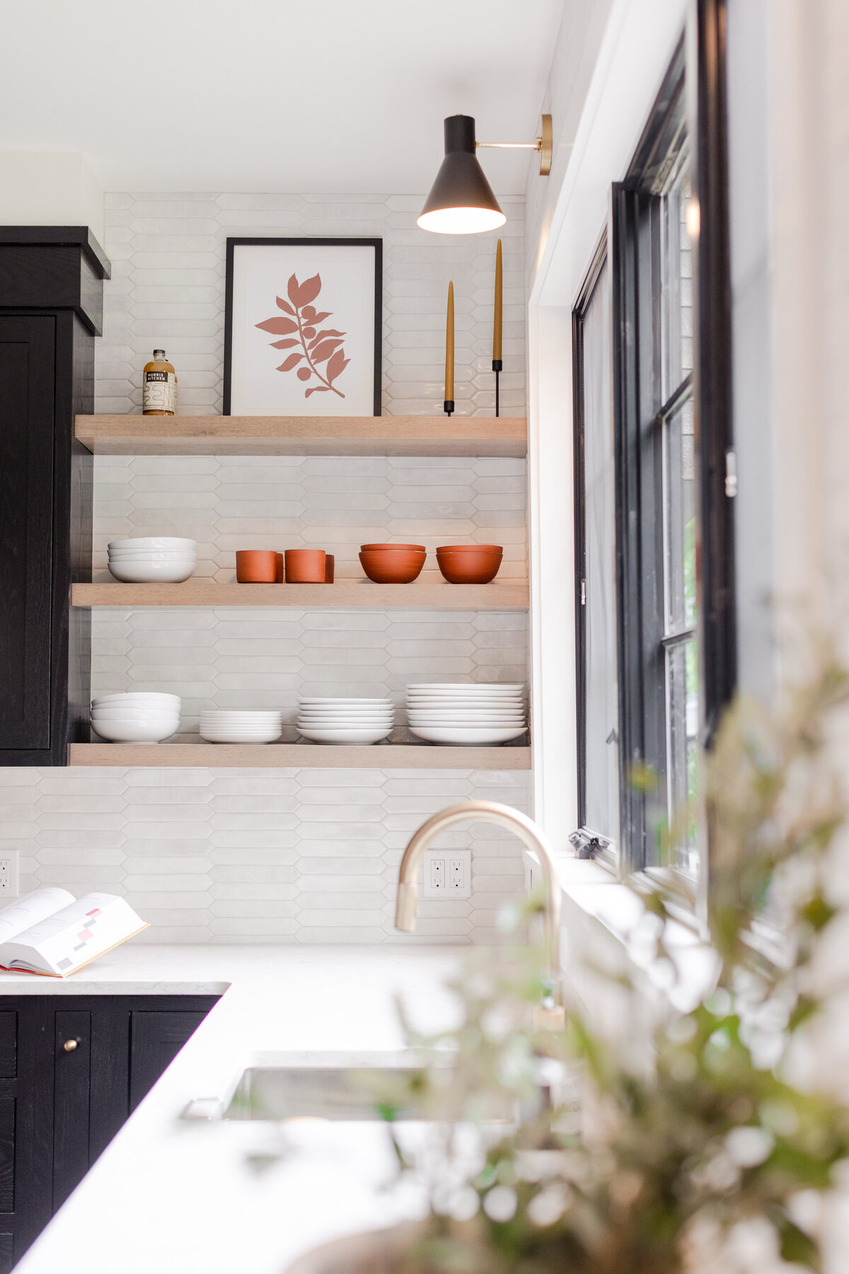 The kitchen decor features wooden floating shelves, black cabinetry, and a white countertop. The custom red oak cabinets have a midnight finish, while the floating shelves are finished in Irish cream. The backsplash is adorned with ceramic picket tiles, complementing the quartz countertop.