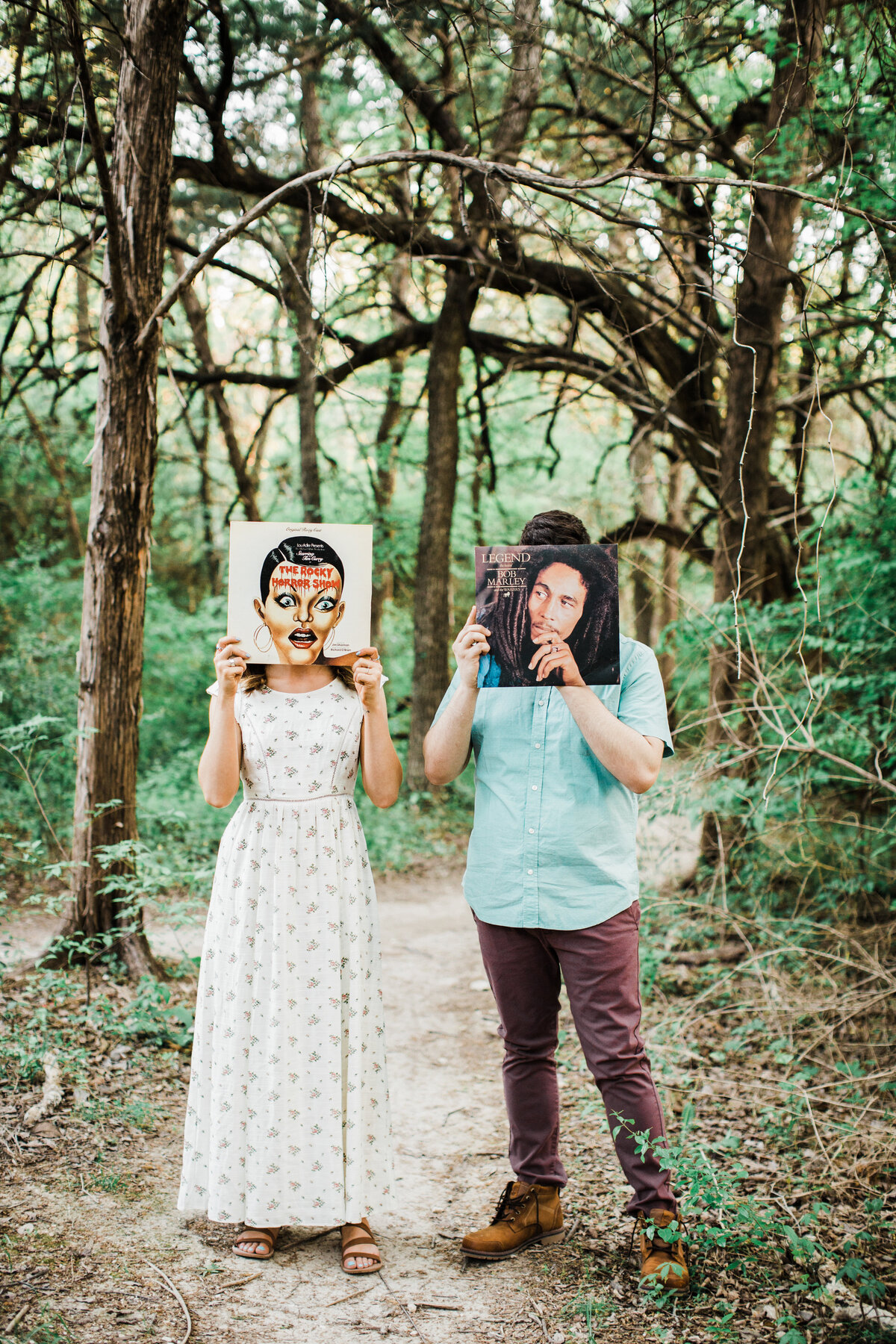 A couple posing in the woods while holding up vinyl album covers in front of their faces during their engagement session in Dallas, Texas. The woman on the left is wearing a long white dress covered in small flowers and is holding up the soundtrack to "The Rocky Horror Picture Show" in front of her face. The man on the right is wearing a blue dress shirt and maroon pants and is holding up Bob Marley's "Legend" in front of his face.