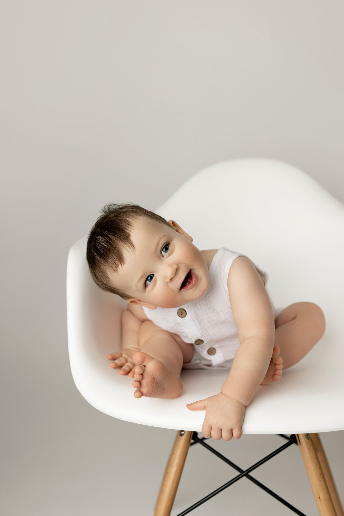 Baby boy in white romper sitting in white eames-style chair. Baby is looking off to the side and smiling. Photo taken in London, Ontario baby photo studio.