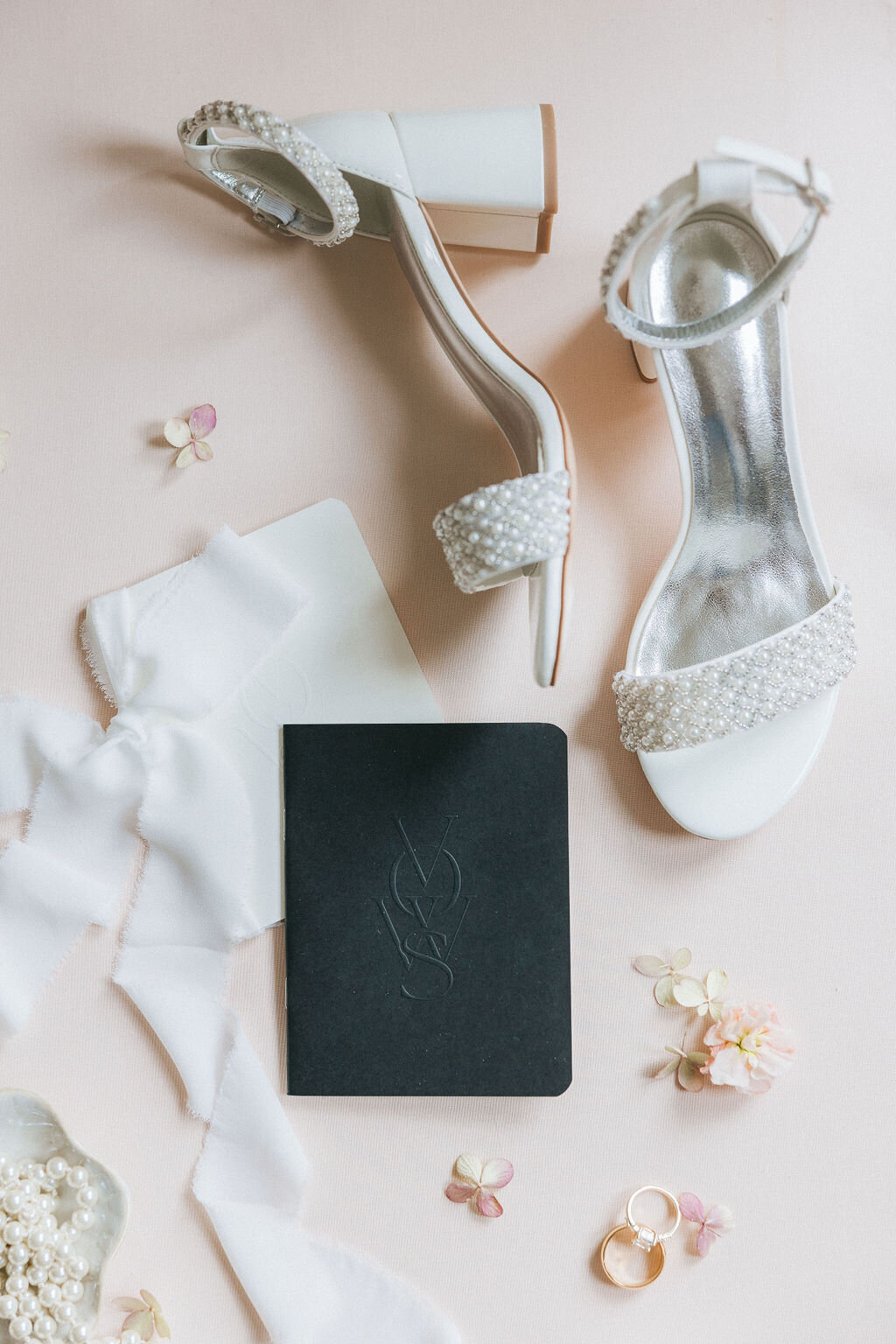 Pearl-embellished bridal heels, black debossed vow book, and silk ribbon styled on a blush backdrop—detail from an Admiral House wedding in Charleston, captured by luxury wedding photographer Amia Marcell