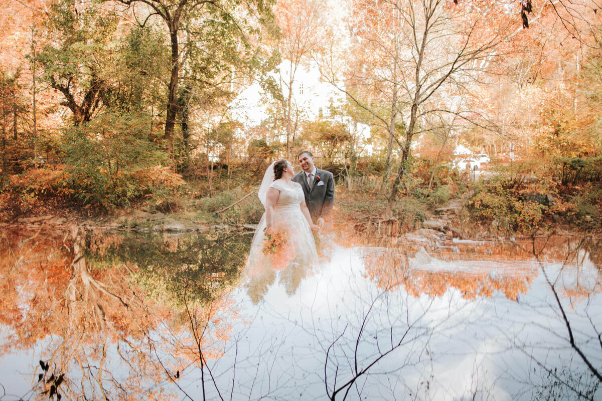 A bride and groom share a joyful moment by a tranquil pond surrounded by vibrant autumn foliage at Valley Green Inn, Philadelphia.