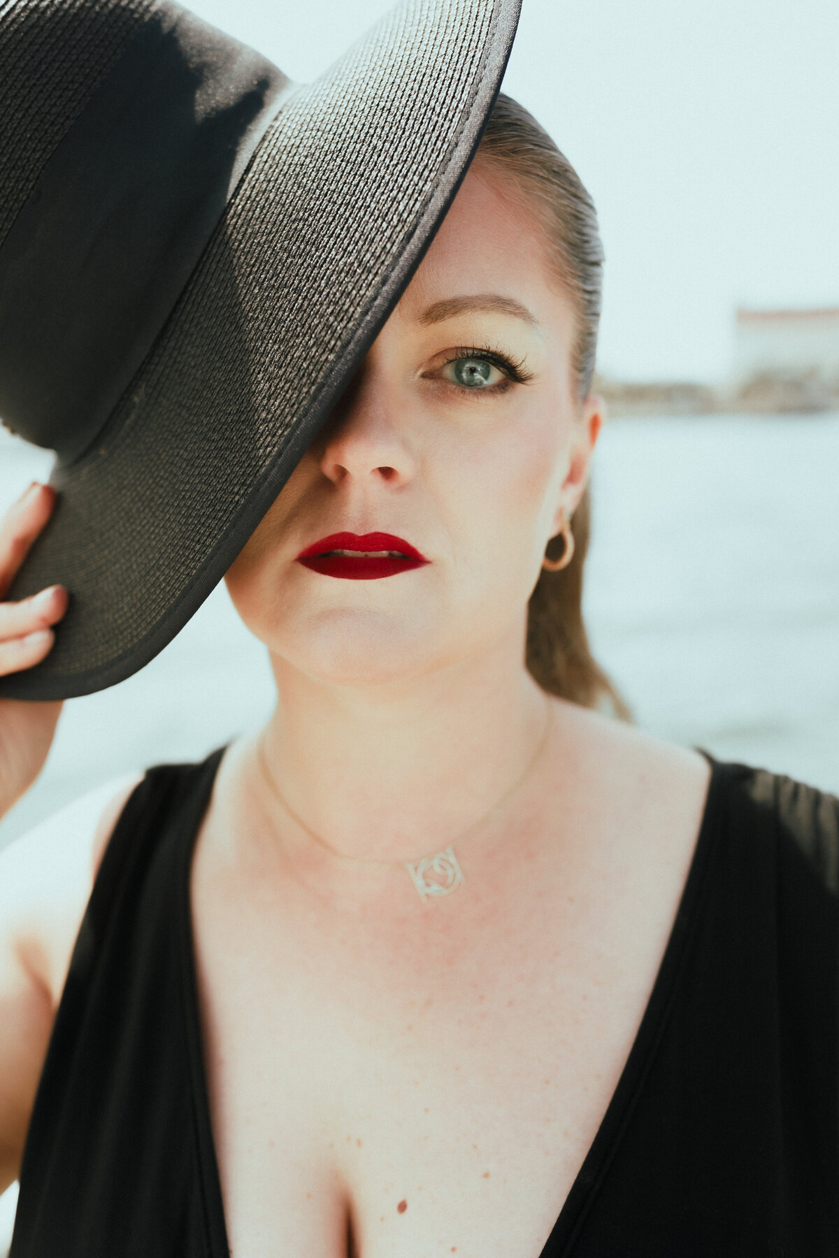 Woman in bold makeup and black outfit holds a wide-brimmed hat over one eye while standing on a boat with coastal backdrop.