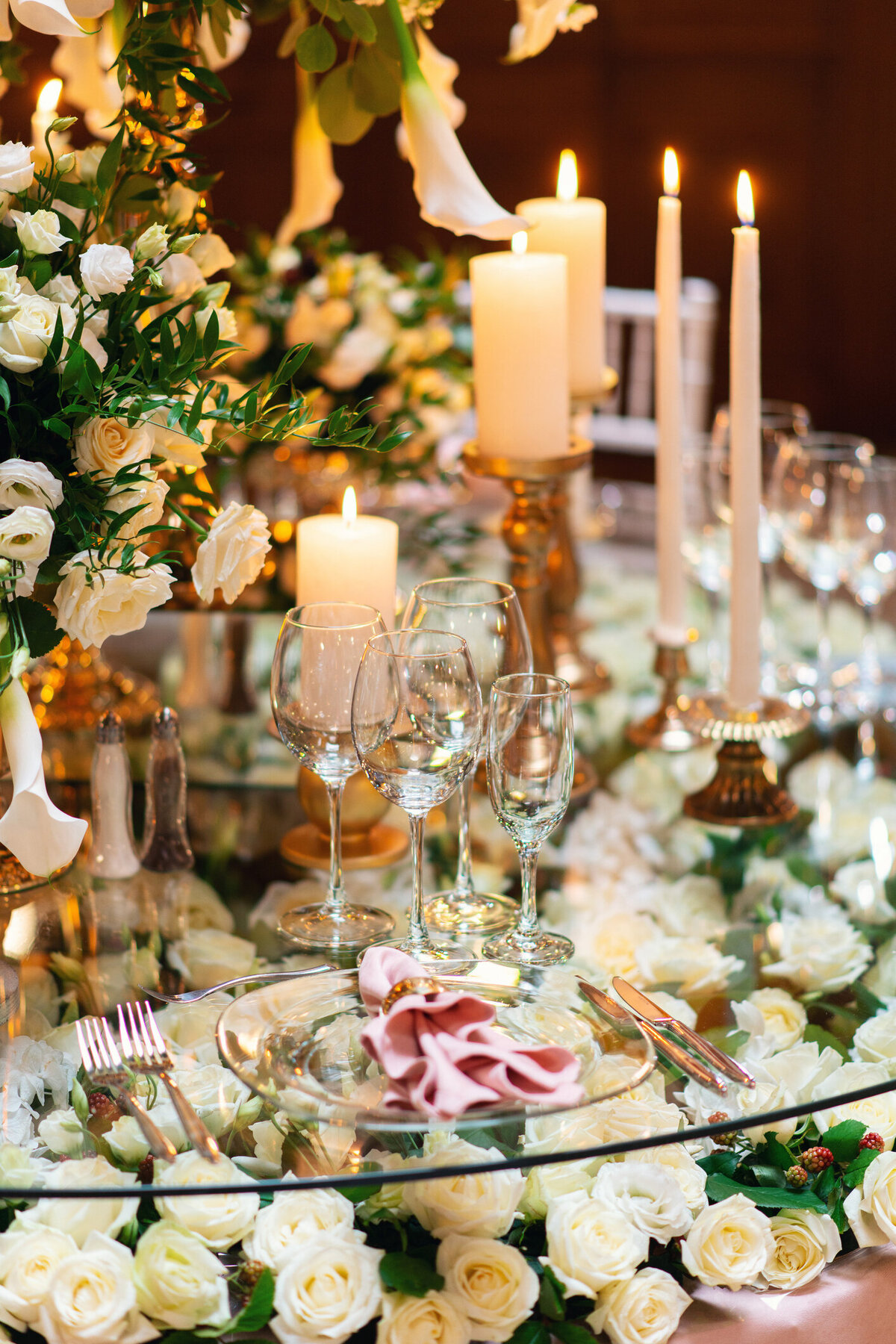 An opulent wedding tablescape adorned with lush roses, glowing candles, and elegant glassware. The rich floral arrangements and warm candlelight create a romantic and sophisticated ambiance, perfect for a timeless and luxurious wedding reception."