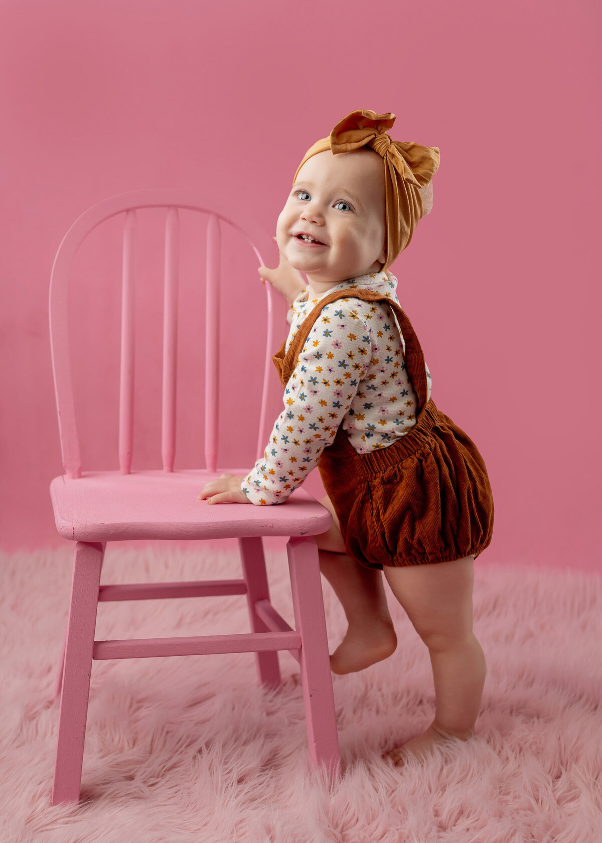 A little girl wearing an orange jumper smiles while she stands next to a pink chair