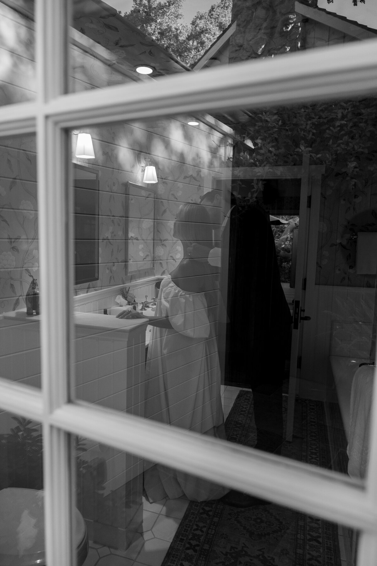 Black‑and‑white photo looking through a window to see a groom standing behind the bride on the other side, tenderly holding her hair and peeking in to kiss her cheek.