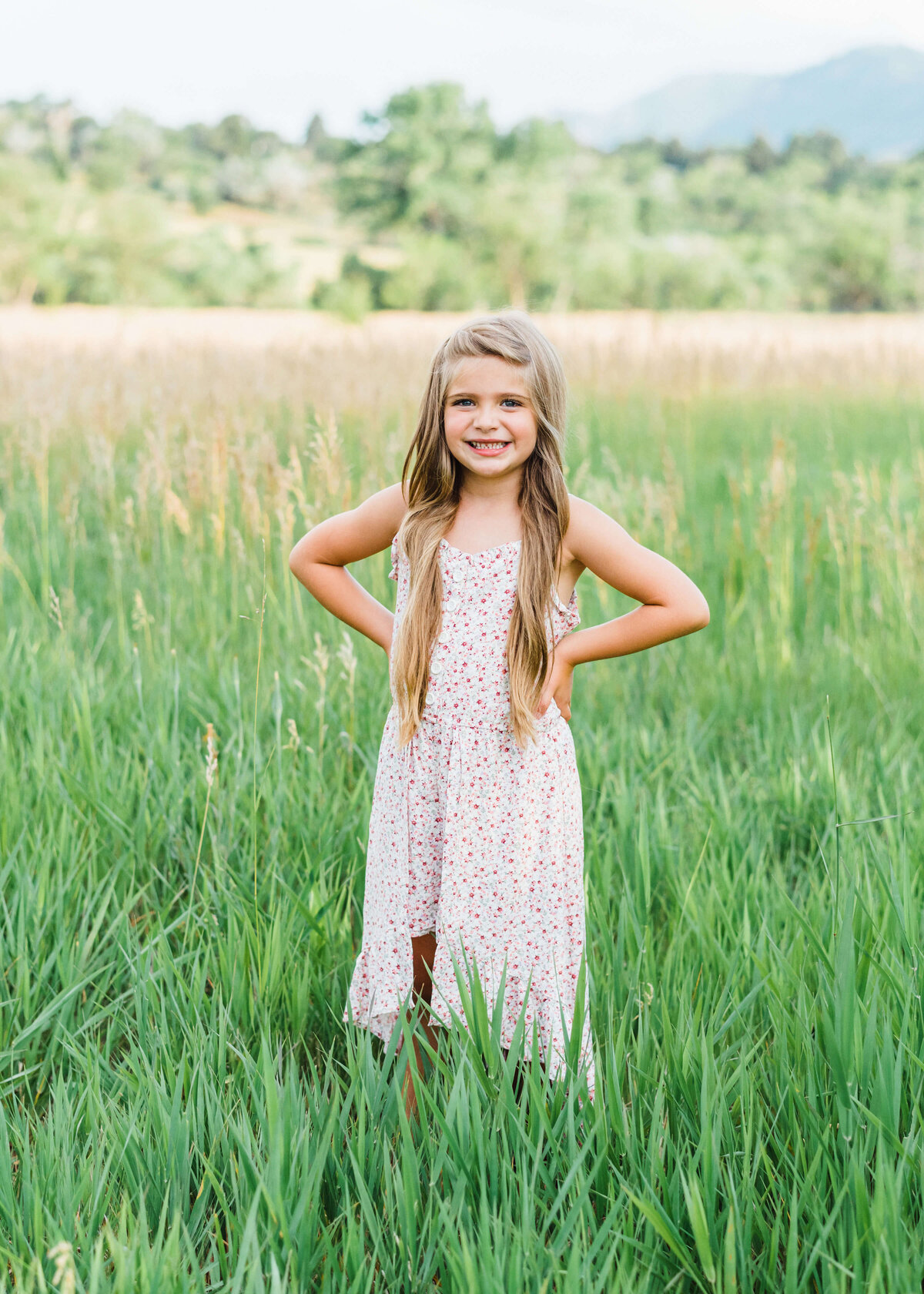 An adorable, blonde young girl stands in a field and smiles at the camera