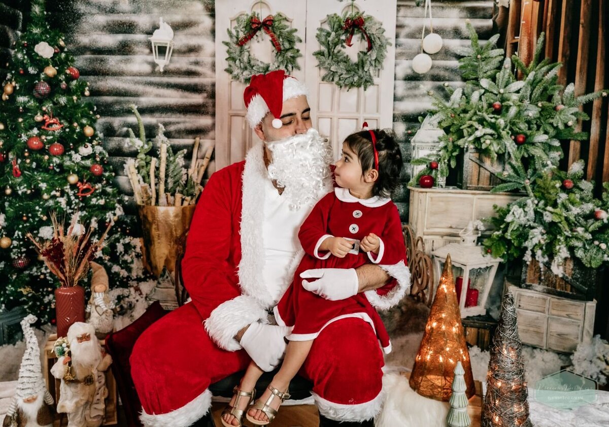 Toddler dressed in a red Santa outfit sitting on Santa Claus’s lap in a holiday-themed studio.